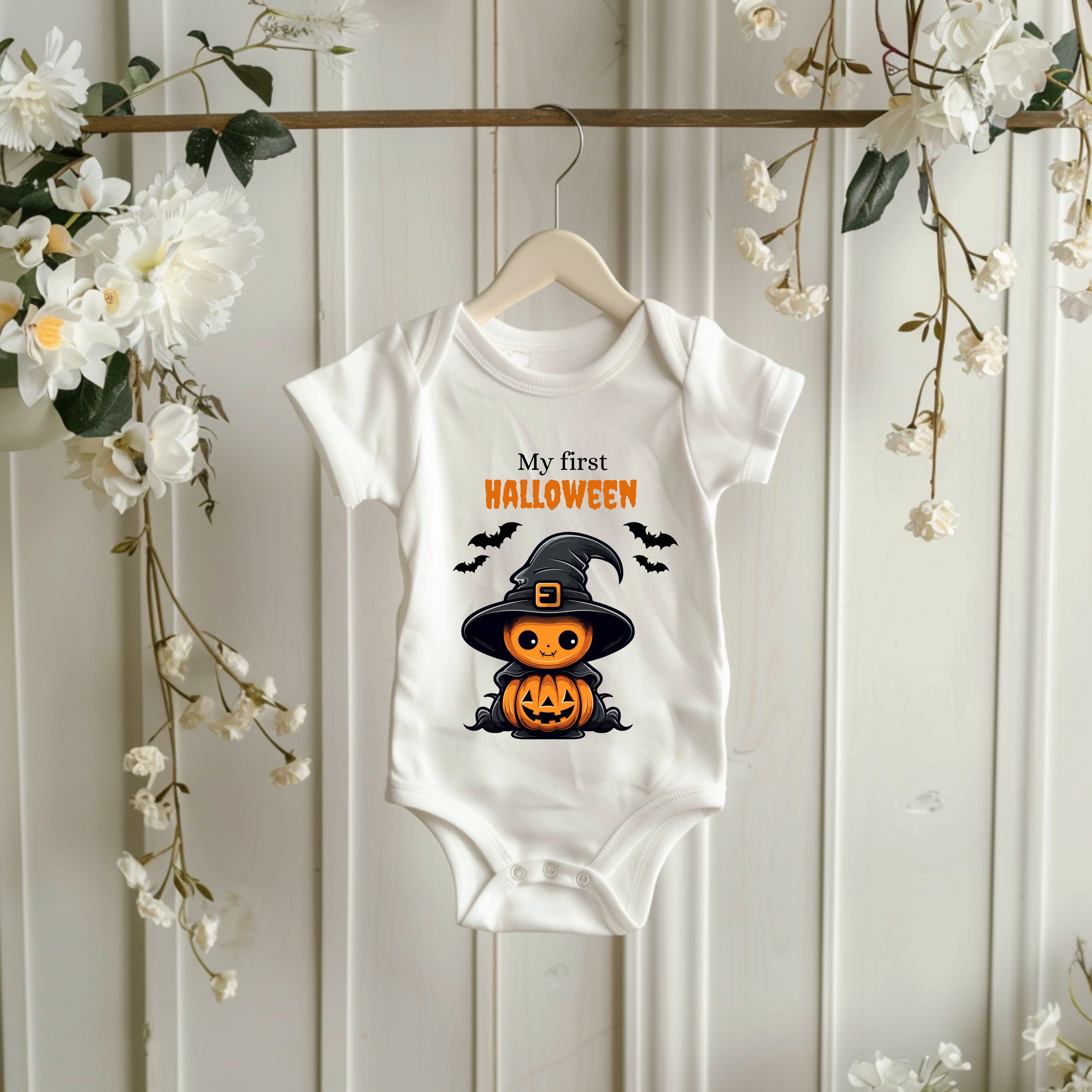 Weißer Baby-Strampler mit Halloween-Motiv: niedlicher Kürbis im Hexenkostüm mit Hut und Umhang, umgeben von Fledermäusen. Darüber der Schriftzug ‚My First Halloween‘.