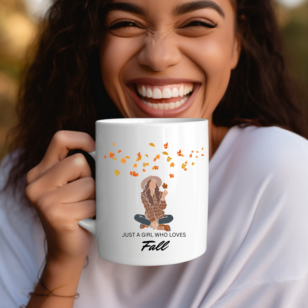 Weiße Keramiktasse mit Illustration einer stilvoll gekleideten Frau im herbstlichen Outfit, umgeben von fallenden orange-, rot- und braunfarbenen Blättern. Darunter der verspielte Schriftzug ‚Just a girl who loves fall‘.
