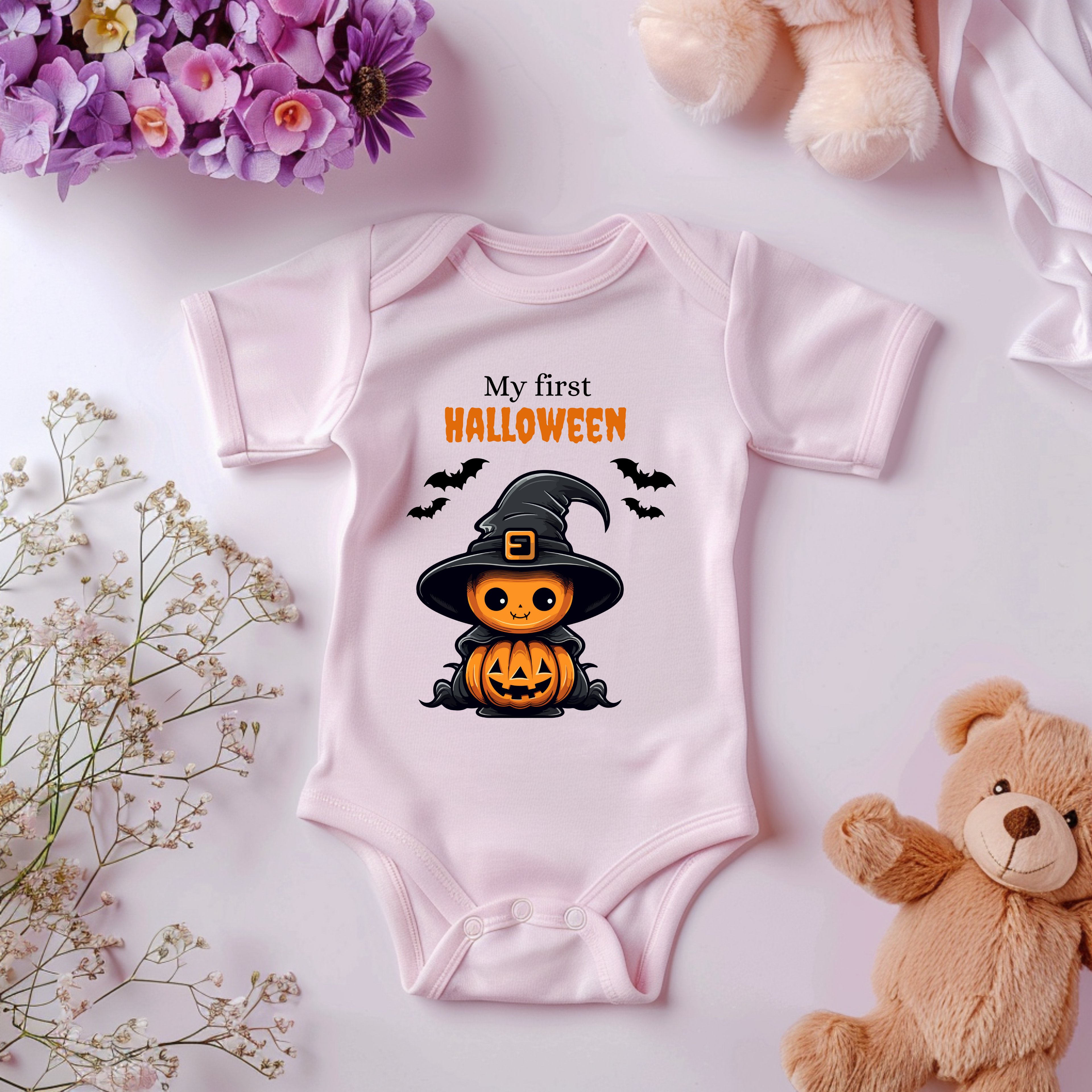 Rosa Baby-Strampler mit Halloween-Motiv: niedlicher Kürbis im Hexenkostüm mit Hut und Umhang, umgeben von Fledermäusen. Darüber der Schriftzug ‚My First Halloween‘.