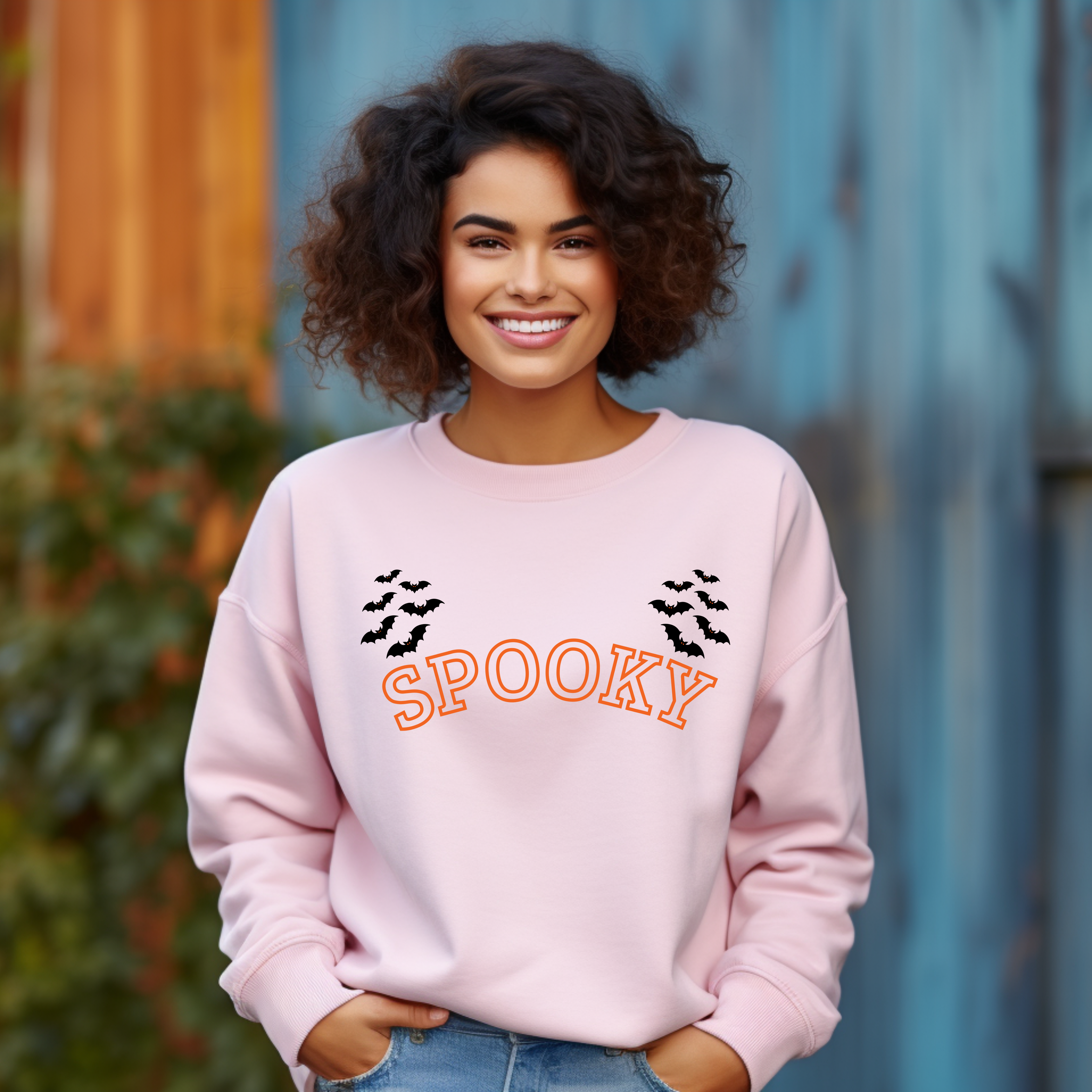 Rosa Halloween-Sweatshirt mit großem orangefarbenem Schriftzug ‚Spooky‘ in der Mitte, umrahmt von mehreren flatternden schwarzen Fledermäusen. Minimalistisches, aber wirkungsvolles Design mit typischem Halloween-Flair.
